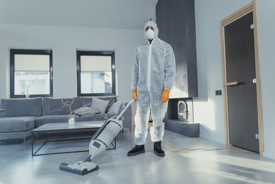 A commercial vacuum cleaner with a metal body, blue accents, and black hoses is positioned on a wooden floor in a residential room. The vacuum hose extends across the floor, ending with a wide cleanin
