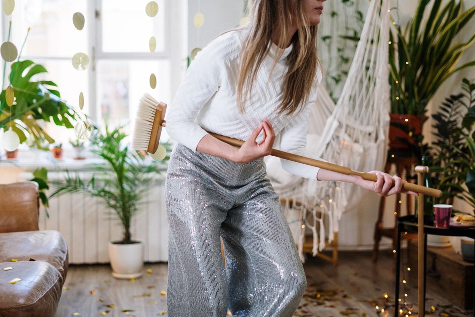 A woman dressed in a white top and silver sequined trousers is engaged in a cleaning activity inside a well-lit living room with large windows. She is holding a broom with both hands, seemingly sweeping the wooden floor which appears clean and shiny. The room features various houseplants in white pots, a leather sofa, and decorative elements like a hammock and string lights, creating a cozy atmosphere. Visible surfaces include the wooden floor, a side table, and a glass on the table. The natural lighting highlights the cleanliness and orderliness of the space. This scene illustrates domestic deep cleaning and surface sanitisation, aligning with professional cleaning services offered by Deep Cleaning Fulham, as referenced in the page 'Parsons Green flat deep cleaning before and after' on deepcleaningfulham.co.uk.