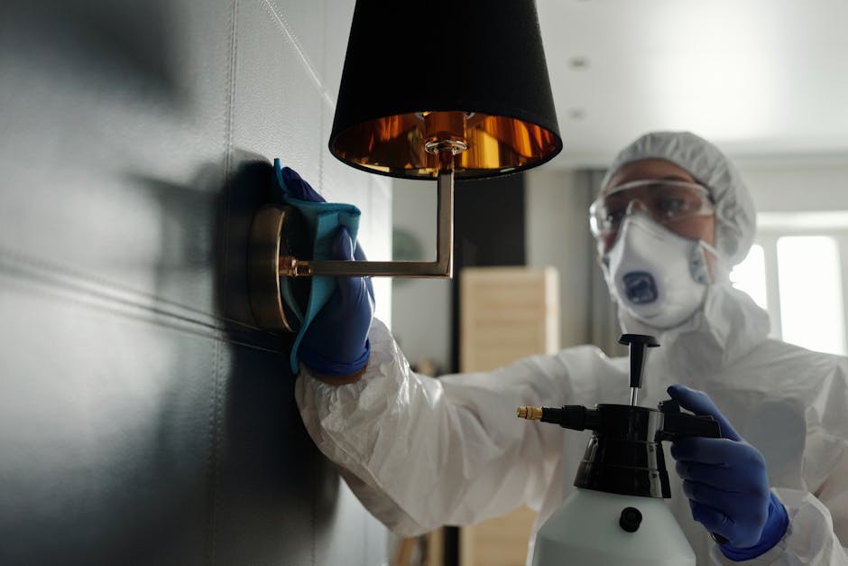 A professional cleaner from Deep Cleaning Fulham is kneeling on a wooden floor in a residential room, wearing protective white coveralls, a mask, goggles, and blue gloves. The cleaner is using a cloth