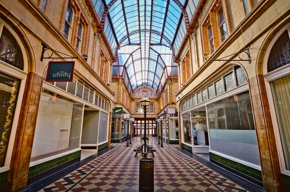 Interior view of a shopping arcade featuring a high, curved glass roof allowing natural light to illuminate the polished tile flooring with a geometric pattern. The arcade is surrounded by storefronts with large glass windows, some displaying products or signage. A central lamppost and benches are situated along the corridor, with a clean and well-maintained appearance. The surfaces, including tiles, glass, and decorative archways, appear spotless and reflective, indicating recent or ongoing deep cleaning and surface sanitisation efforts by [COMPANY_NAME]. The overall scene showcases a spacious and hygienic environment suitable for domestic or commercial cleaning reference, aligned with the page about deep cleaning near Fulham Broadway SW6.
